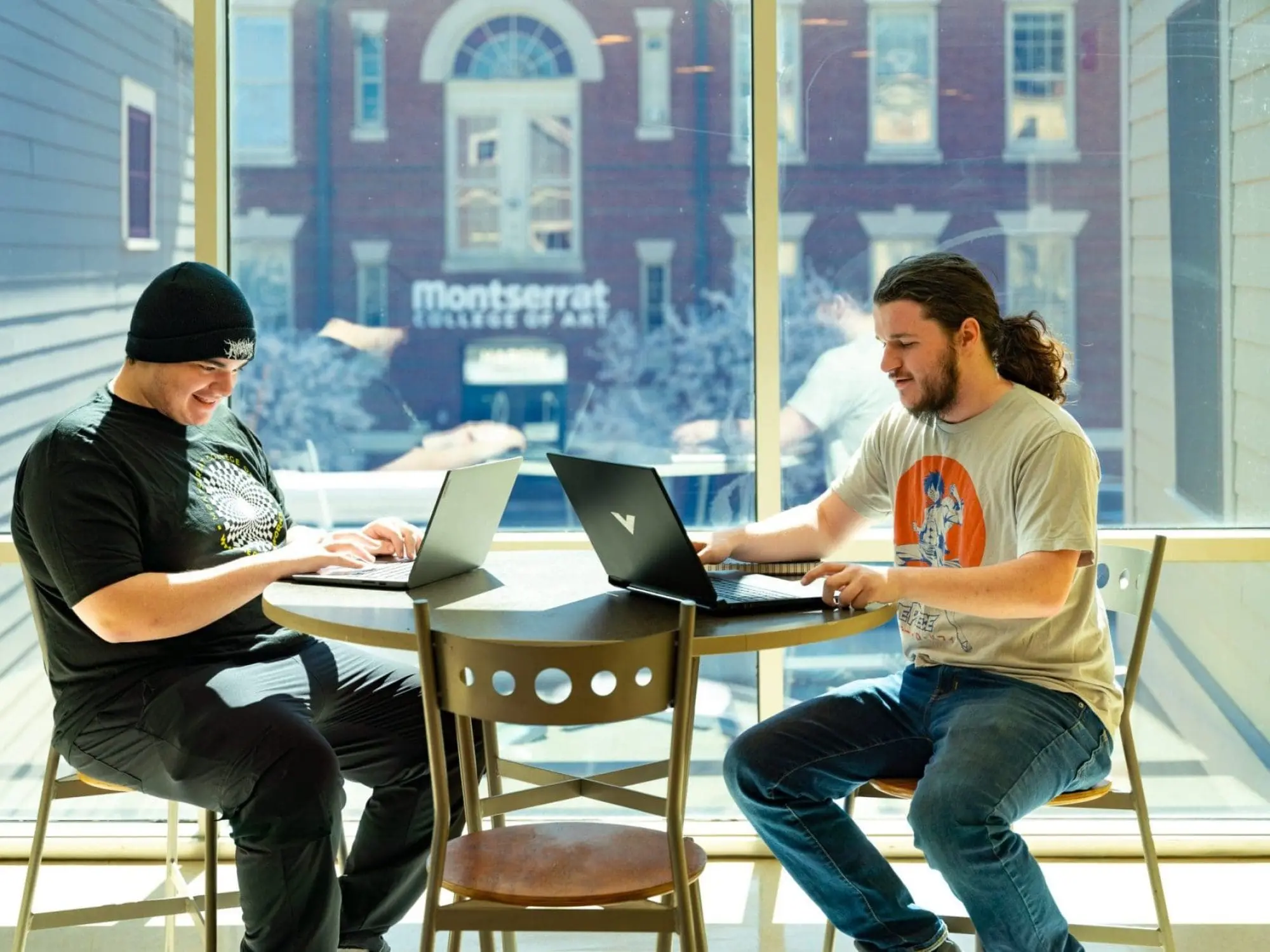 two students sitting in a window working on their laptops