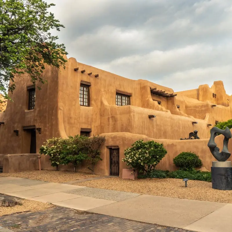 Pueblo Revival Style building with earth tone color, rounded corners and battered walls under dramatic cloudy sky, side, New Mexico Museum of Art, Santa Fe, New Mexico